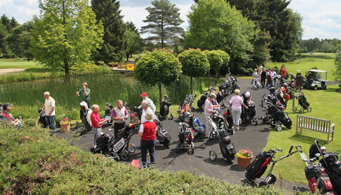 Eine Gruppe von Golfern versammelt sich mit Golf-Trolleys auf einem Weg in einem Parkbereich.