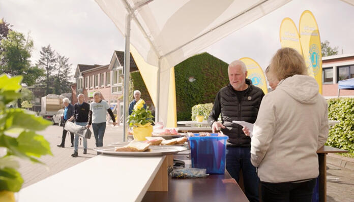 Menschen an einem Verkaufs- oder Empfangsstand im Freien vor Gebäuden, mit Kuchen auf Tischen.